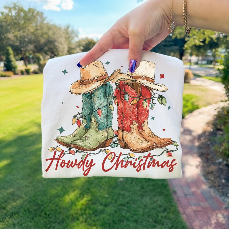 May include: White t-shirt featuring a watercolor illustration of cowboy boots adorned with Christmas lights and cowboy hats. The text "Howdy Christmas" is written in red script below the boots.