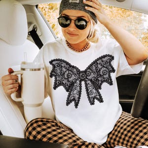 May include: White t-shirt featuring a large black lace bow design. The person in the image is wearing a gray beanie, sunglasses, and a gold chain necklace. They are holding a white tumbler with a straw. The person is wearing plaid pants and black sneakers.
