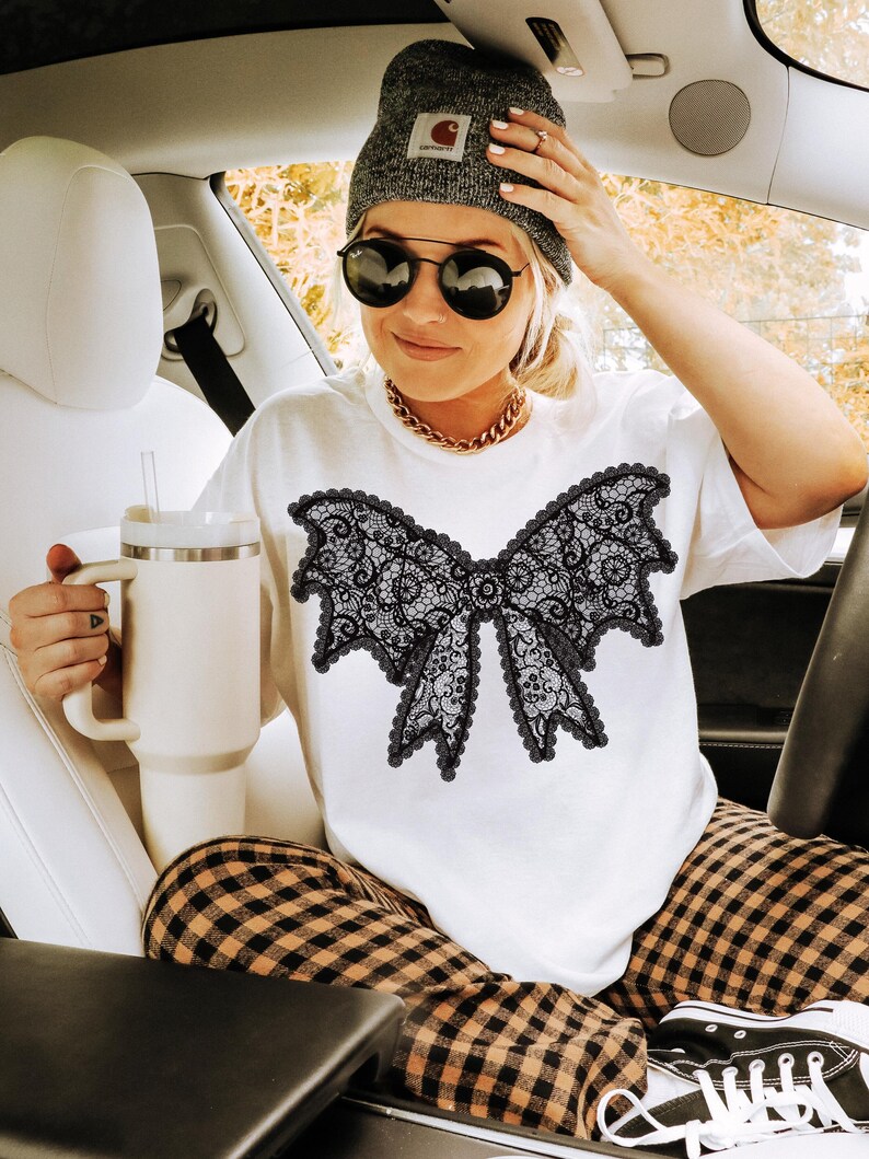 May include: White t-shirt featuring a large black lace bow design. The person in the image is wearing a gray beanie, sunglasses, and a gold chain necklace. They are holding a white tumbler with a straw. The person is wearing plaid pants and black sneakers.