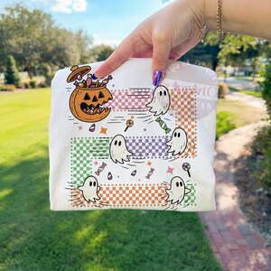 May include: White t-shirt featuring a Halloween-themed design. The graphic includes a jack-o'-lantern filled with candy, cartoon ghosts, and various candies. The design incorporates checkered patterns in green, purple, and orange.