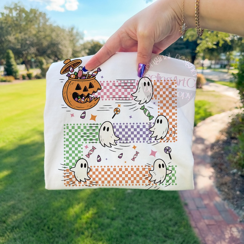 May include: White t-shirt featuring a Halloween-themed design. The graphic includes a jack-o'-lantern filled with candy, cartoon ghosts, and various candies. The design incorporates checkered patterns in green, purple, and orange.