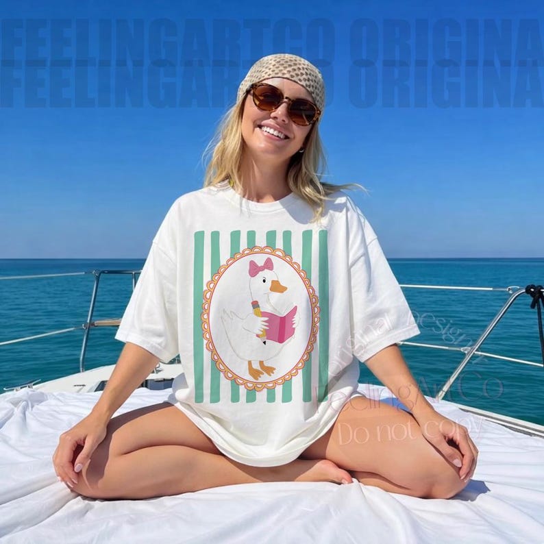 May include: White t-shirt with a cartoon goose reading a pink book, set within a decorative oval frame. The shirt has vertical mint green stripes and is worn by a person on a boat. The background is the ocean and a clear blue sky.