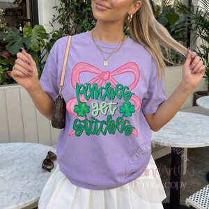 May include: A light purple short-sleeve t-shirt featuring a large pink bow graphic and the green text "pinches get stitches" with two green shamrock icons. The shirt is worn with a brown shoulder bag featuring a dark brown monogram pattern.