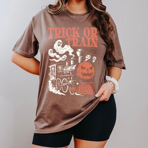 May include: Brown t-shirt with the text "TRICK OR TRAIN" in orange. The graphic features a Halloween-themed train with a jack-o'-lantern, ghosts, and skulls. The shirt is a short-sleeved, oversized style.