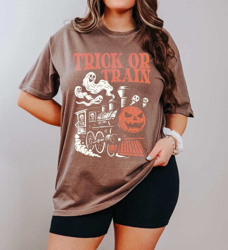 May include: Brown t-shirt with the text "TRICK OR TRAIN" in orange. The graphic features a Halloween-themed train with a jack-o'-lantern, ghosts, and skulls. The shirt is a short-sleeved, oversized style.
