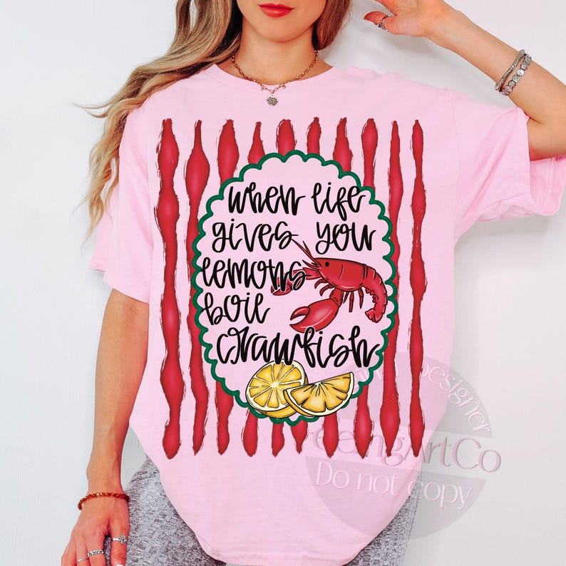 K&ouml;nnte beinhalten: Hellrosa T-Shirt mit dem Text "When life gives you lemons, boil crawfish". Die Grafik zeigt einen roten Krebs und Zitronenscheiben. Das Shirt hat rote vertikale Streifen.