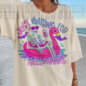 Peut inclure: T-shirt cr&egrave;me avec un squelette portant des lunettes de soleil, se pr&eacute;lassant sur un flotteur flamant rose avec une boisson. Le texte "JUST WAITING FOR HALLOWEEN" est en violet et sarcelle, avec des chauves-souris. Un design amusant et effrayant.