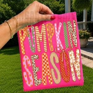 May include: A bright pink t-shirt featuring the text PUMPKIN SEASON in large, multi-colored letters. Each letter displays a unique pattern, including leopard print, orange and white checkers, pink flowers, and stars, with two pink bows.