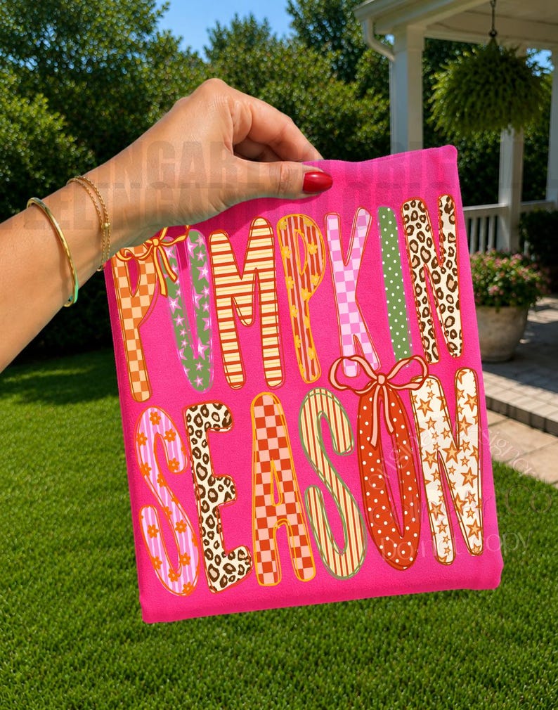 May include: A bright pink t-shirt featuring the text PUMPKIN SEASON in large, multi-colored letters. Each letter displays a unique pattern, including leopard print, orange and white checkers, pink flowers, and stars, with two pink bows.