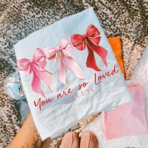 Pu&ograve; includere: Una maglietta azzurra con tre fiocchi ad acquerello nei toni del rosa e del rosso con dettagli dorati. Il testo "You are so Loved" e "John 3:16" &egrave; stampato in rosso.