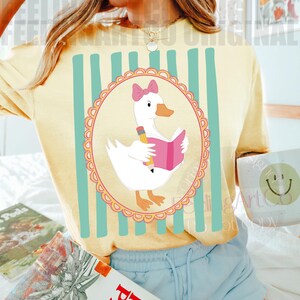 May include: Yellow t-shirt with a cartoon goose reading a book, pencil in hand, inside a pink-trimmed oval. The shirt has vertical teal stripes and the text "FEELING ART ORIGINAL".