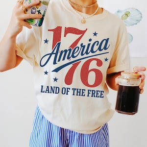 May include: A cream-coloured t-shirt with the text "1776 America Land of the Free" in red and blue. The person is wearing blue and white striped shorts and holding a phone case with a colourful design and a glass with a drink.
