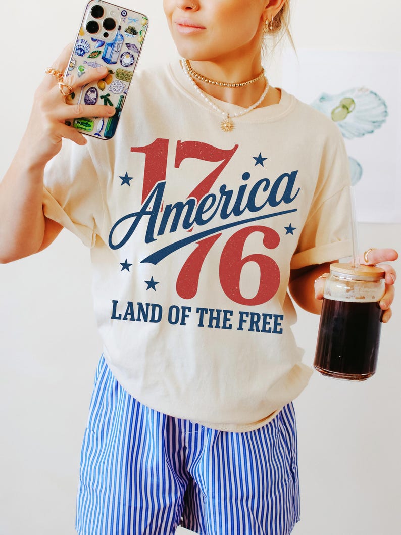 May include: A cream-coloured t-shirt with the text "1776 America Land of the Free" in red and blue. The person is wearing blue and white striped shorts and holding a phone case with a colourful design and a glass with a drink.