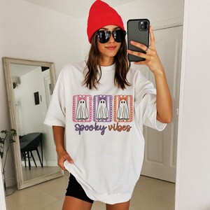 May include: White t-shirt with a graphic of three cartoon ghosts in rectangular frames, each with a bow. The text "Spooky Vibes" is below the ghosts. The shirt is worn with a red beanie and sunglasses.