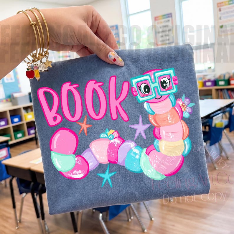May include: A gray t-shirt featuring a colorful cartoon worm wearing teal glasses, with the word BOOK printed in bright pink letters above it. The design includes small star shapes and floral accents in shades of pink, blue, and yellow.