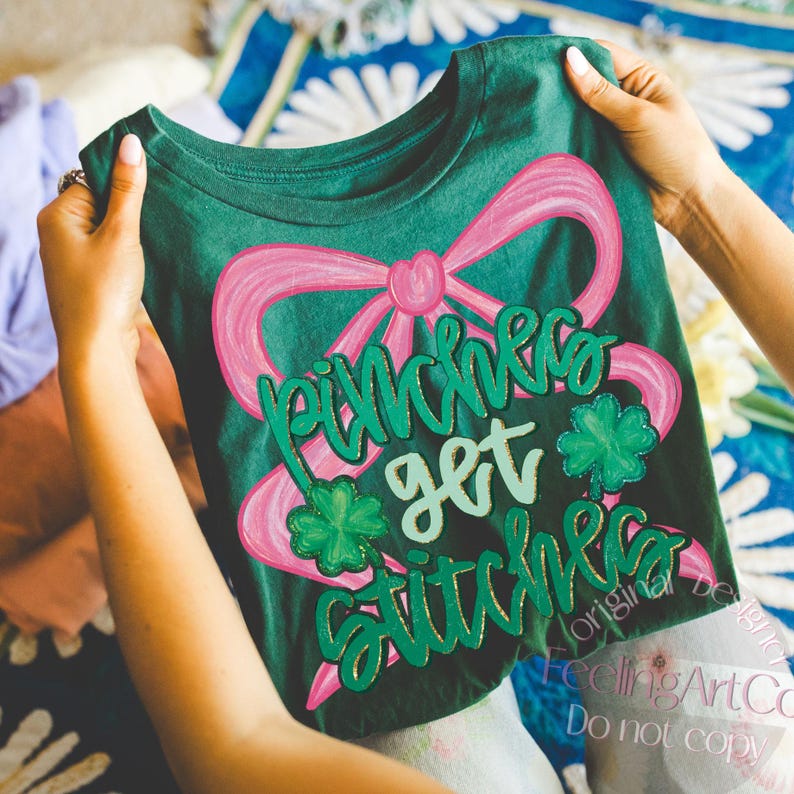 May include: Green t-shirt with a pink bow and the words "Pinches get stitches" in green script. The design includes green shamrocks and gold accents. The shirt is held up by a person.