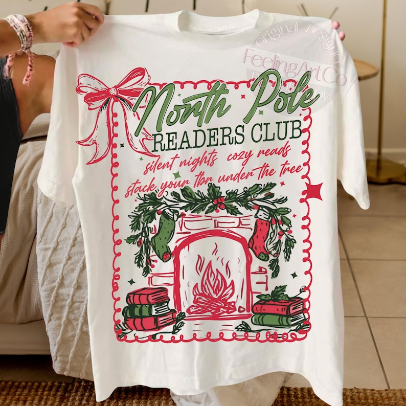 Op de afbeelding: Een wit t-shirt met een feestelijk ontwerp voor de North Pole Readers Club. De afbeelding bevat een open haard, kousen, boeken en de tekst "silent nights cozy reads stack your ton under the tree". Een rode strik en decoratieve rand maken het ontwerp compleet.