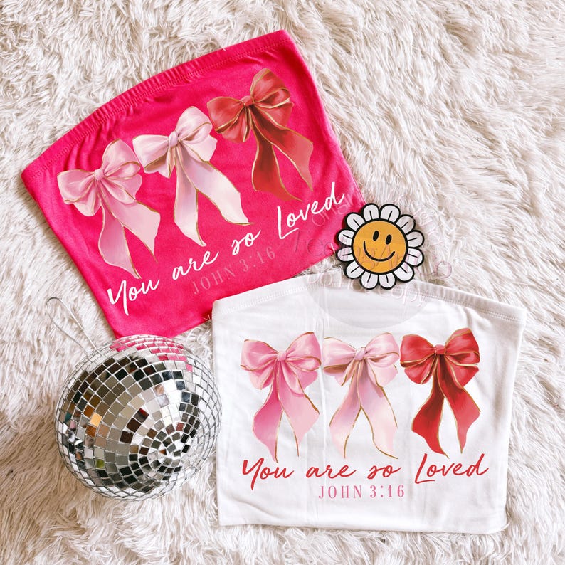 Pu&ograve; includere: Due top: un top a tubo rosa acceso e una maglietta bianca. Entrambi presentano tre fiocchi rosa con dettagli dorati e il testo "You are so Loved" e "John 3:16". Una toppa a forma di fiore con faccina sorridente e una palla da discoteca sono anche nell'immagine.