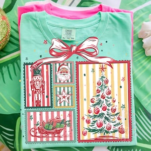 May include: A mint green t-shirt with a Christmas-themed graphic featuring a nutcracker, Santa Claus, a star, a Christmas tree, and a sleigh filled with gifts. The design is framed with red borders and a large red bow. A pink t-shirt is visible underneath.