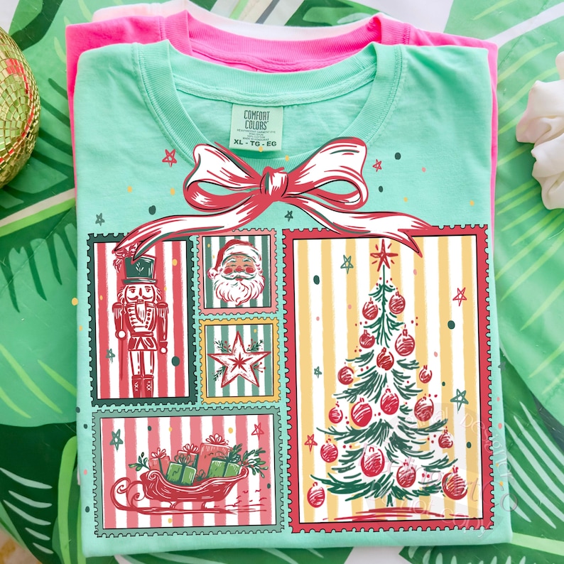 May include: A mint green t-shirt with a Christmas-themed graphic featuring a nutcracker, Santa Claus, a star, a Christmas tree, and a sleigh filled with gifts. The design is framed with red borders and a large red bow. A pink t-shirt is visible underneath.