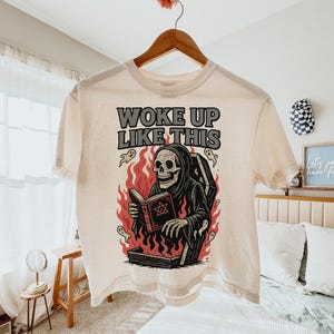 May include: A cream-coloured t-shirt featuring a graphic of a skeleton in a black robe reading a book, emerging from a coffin surrounded by flames and ghosts. The text "WOKE UP LIKE THIS" is printed above the graphic.
