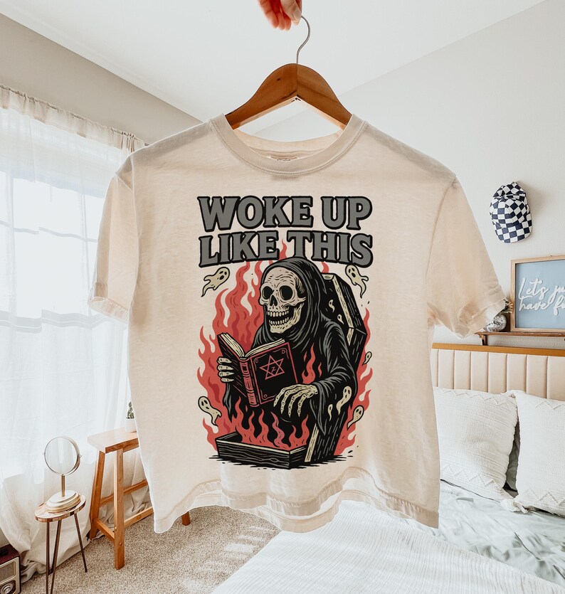 May include: A cream-coloured t-shirt featuring a graphic of a skeleton in a black robe reading a book, emerging from a coffin surrounded by flames and ghosts. The text "WOKE UP LIKE THIS" is printed above the graphic.
