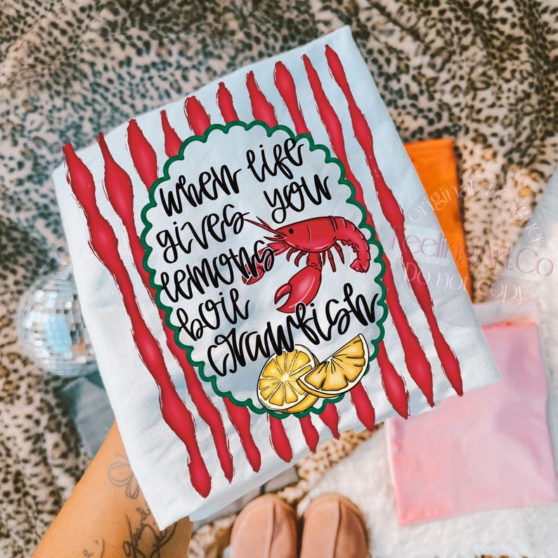 K&ouml;nnte beinhalten: Wei&szlig;es K&uuml;chentuch mit roten vertikalen Streifen und dem Text "When life gives you lemons boil crawfish". Das Design umfasst einen roten Krebs und Zitronenscheiben. Das Handtuch liegt auf einer gemusterten Oberfl&auml;che.