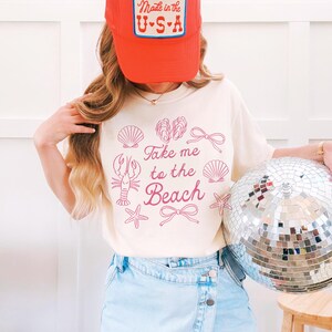May include: A cream-colored t-shirt with the phrase "Take me to the Beach" and beach-themed illustrations. The person is wearing a red trucker hat with "Made in the U.S.A." on it and light blue denim shorts. A silver disco ball is held in the right hand.