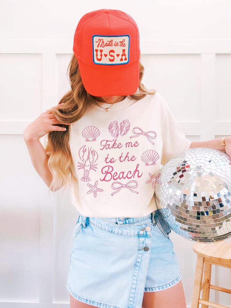 May include: A cream-colored t-shirt with the phrase "Take me to the Beach" and beach-themed illustrations. The person is wearing a red trucker hat with "Made in the U.S.A." on it and light blue denim shorts. A silver disco ball is held in the right hand.