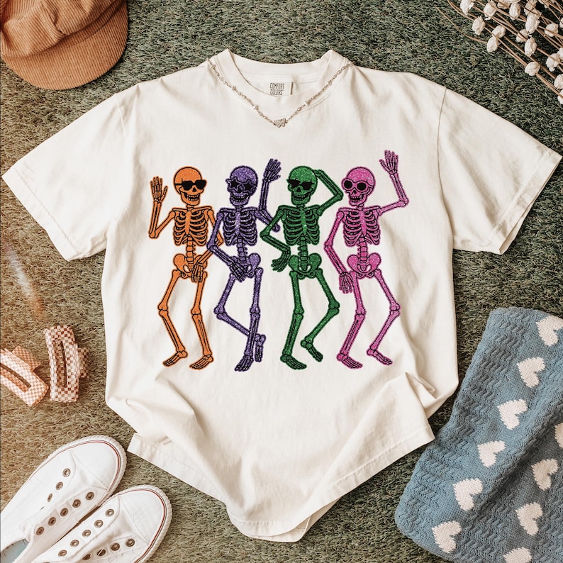 Puede incluir: Camiseta de color crema con un dise&ntilde;o de cuatro esqueletos bailando en naranja, morado, verde y rosa. Cada esqueleto lleva gafas de sol. La camiseta tiene cuello en V y mangas cortas. Se ve un collar plateado.