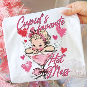 May include: White t-shirt featuring a pink illustration of Cupid in a martini glass, with the text "Cupid's favorite Hot Mess." Pink hearts and a bow accent the design. The shirt is held by a hand.