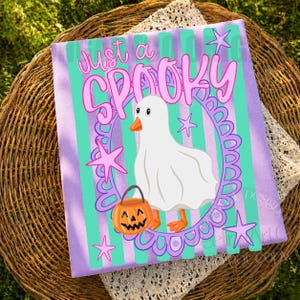 May include: A rectangular fabric print featuring a white ghost-costumed duck holding an orange jack-o'-lantern bucket. The design includes a purple scalloped oval frame, teal vertical stripes, pink stars, and the text Just a SPOOKY.