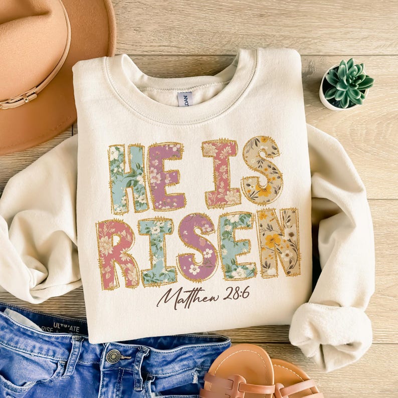 May include: A cream-colored sweatshirt with a floral print design that reads "He is Risen" with the scripture reference "Matthew 28:6" underneath.