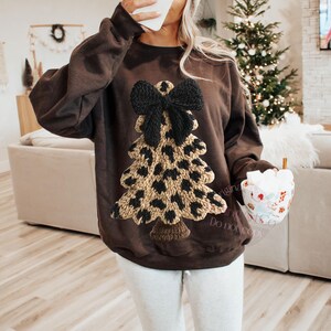 May include: A brown sweatshirt featuring a knitted Christmas tree design with a black bow and leopard print pattern. The tree is made of tan and black yarn. The sweatshirt is a cozy, festive garment.