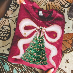 Pu&ograve; includere: Felpa bordeaux con un design ad albero di Natale, un grande fiocco rosa e dettagli dorati. L'albero &egrave; verde con fiocchi rosa e decorazioni dorate. Il design &egrave; centrato sulla parte anteriore della felpa.