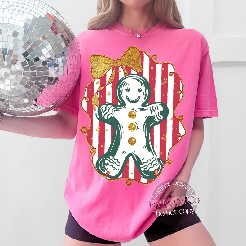 May include: A pink t-shirt featuring a gingerbread man design. The gingerbread man is outlined in green with gold button accents, set against a red and white striped background. A gold glitter bow tops the design. The shirt has the text "FeelingArtCo Do not copy".