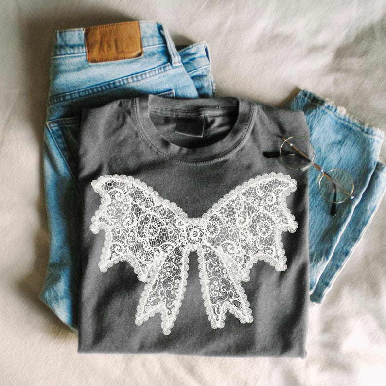 May include: A gray t-shirt featuring a large white lace bow design. The shirt is paired with light blue denim jeans and a pair of eyeglasses. The items are arranged on a neutral-colored surface.