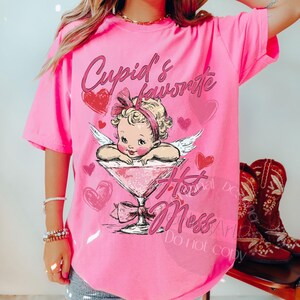 May include: A bright pink t-shirt featuring a graphic of Cupid in a martini glass, with the text "Cupid's Favorite Hot Mess." The design includes hearts and angel wings, perfect for Valentine's Day or a fun, flirty look.