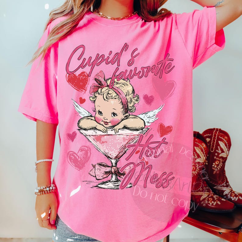 May include: A bright pink t-shirt featuring a graphic of Cupid in a martini glass, with the text "Cupid's Favorite Hot Mess." The design includes hearts and angel wings, perfect for Valentine's Day or a fun, flirty look.