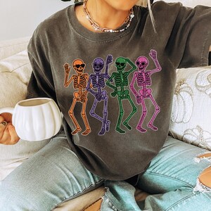 Puede incluir: Camiseta gris oscuro con un dise&ntilde;o de cuatro esqueletos bailando en naranja, morado, verde y rosa. Cada esqueleto lleva gafas de sol. La camiseta es de estilo casual, perfecta para Halloween o un look divertido y original.