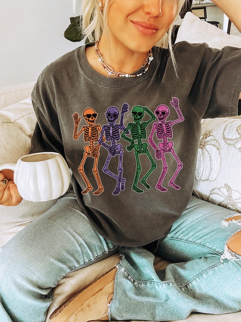 Puede incluir: Camiseta gris oscuro con un dise&ntilde;o de cuatro esqueletos bailando en naranja, morado, verde y rosa. Cada esqueleto lleva gafas de sol. La camiseta es de estilo casual, perfecta para Halloween o un look divertido y original.
