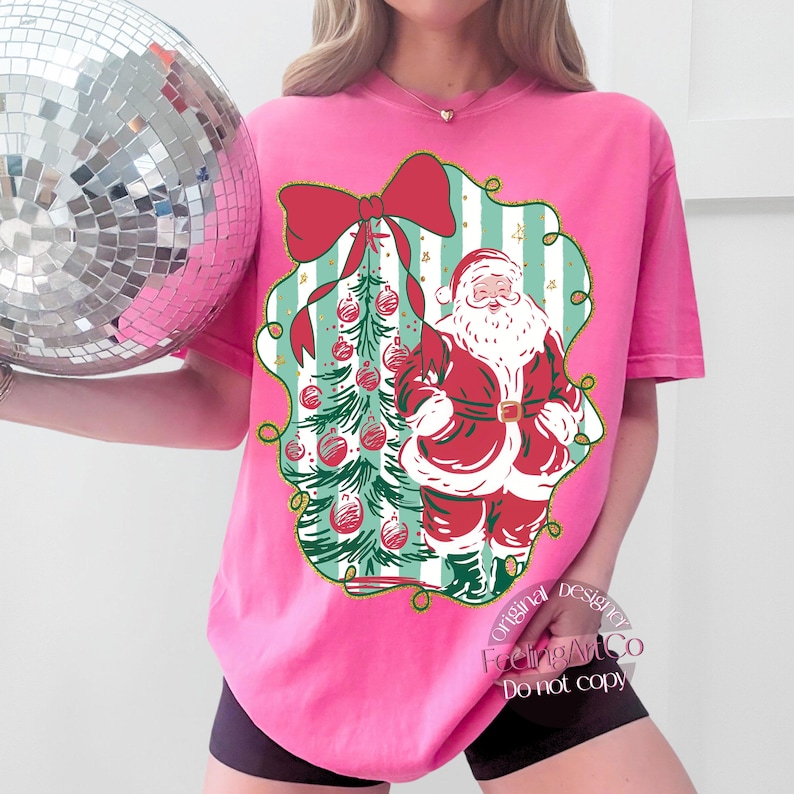 May include: A pink t-shirt featuring a Christmas design. The graphic includes a red bow, a Christmas tree with red ornaments, and Santa Claus. The design is framed by a green and gold border. The text "FeelingArtCo Do not copy" is visible.
