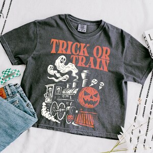 May include: Dark grey t-shirt with the text "TRICK OR TRAIN" in orange. The shirt features a Halloween-themed graphic of a train with a jack-o'-lantern, ghosts, and skulls. A pair of blue jeans and a green and white hair clip are next to the shirt.