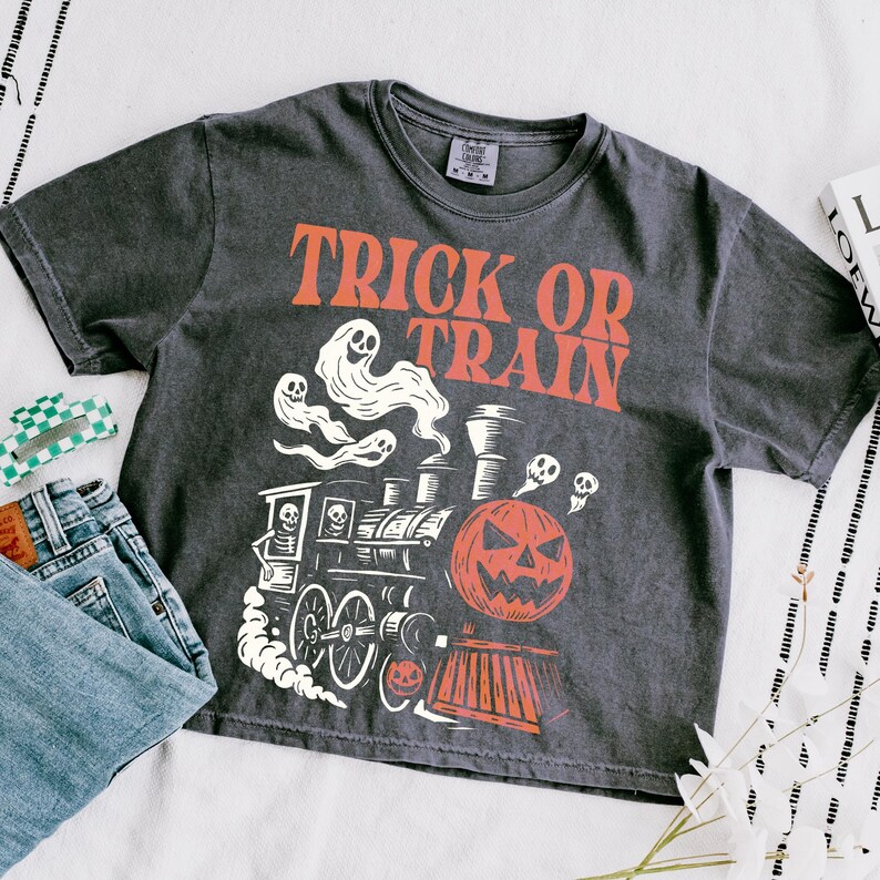 May include: Dark grey t-shirt with the text "TRICK OR TRAIN" in orange. The shirt features a Halloween-themed graphic of a train with a jack-o'-lantern, ghosts, and skulls. A pair of blue jeans and a green and white hair clip are next to the shirt.