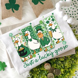 May include: White St. Patrick's Day kitchen towel featuring four cartoon geese in green outfits, each with a different St. Patrick's Day theme. The towel has the text "Just a lucky goose" in green script.