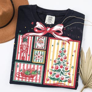 May include: A navy blue sweatshirt features a festive Christmas design. The graphic includes a nutcracker, Santa Claus, a star, a sleigh with gifts, and a Christmas tree. A red bow tops the design. The shirt has the words "Original Feeling Art" on it.
