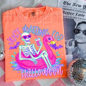 Peut inclure: T-shirt orange Comfort Colors avec un squelette portant des lunettes de soleil sur un flotteur flamant rose, tenant une boisson. Le texte dit "Just Waiting For Halloween". Des chauves-souris violettes et des d&eacute;tails d'eau compl&egrave;tent le design.
