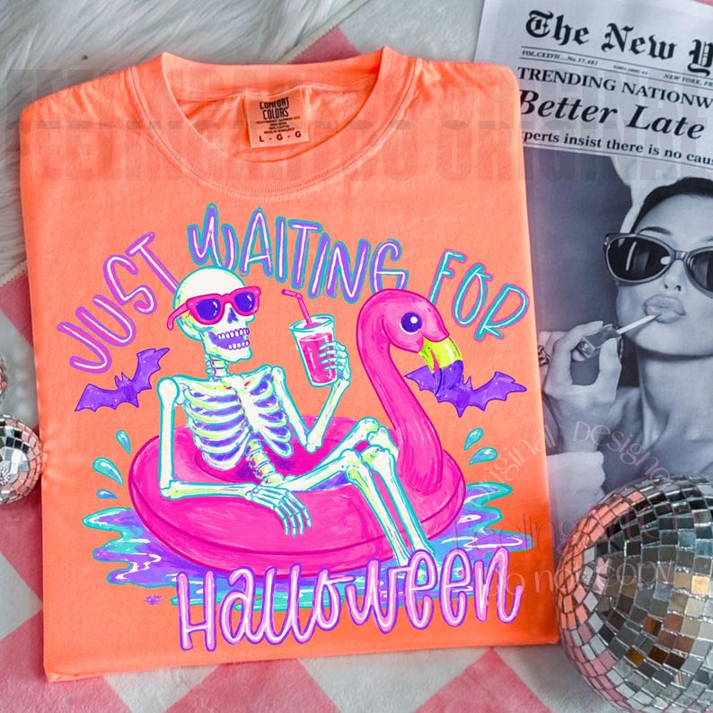 Peut inclure: T-shirt orange Comfort Colors avec un squelette portant des lunettes de soleil sur un flotteur flamant rose, tenant une boisson. Le texte dit "Just Waiting For Halloween". Des chauves-souris violettes et des d&eacute;tails d'eau compl&egrave;tent le design.