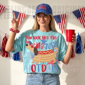 May include: Light blue t-shirt with a graphic of a hotdog dachshund wearing a patriotic hat and sunglasses. The shirt has the text "You look like the 4th of July Hotdog". A blue "USA" baseball cap is worn.