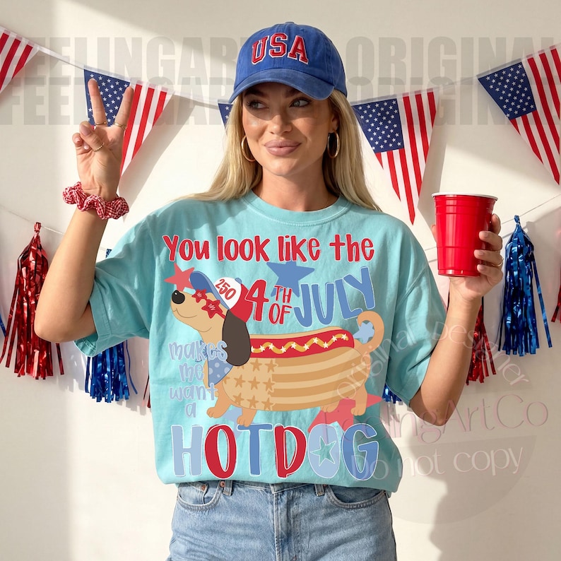 May include: Light blue t-shirt with a graphic of a hotdog dachshund wearing a patriotic hat and sunglasses. The shirt has the text "You look like the 4th of July Hotdog". A blue "USA" baseball cap is worn.